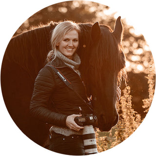 Profile photo of equine photographer Anna Archinger standing with her camera and a Friesian horse at sunset.